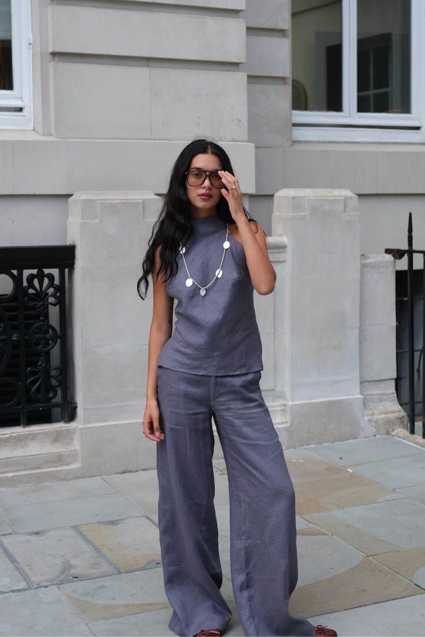 Confident woman walking through the city in a slate blue halterneck linen top with a high neckline and side lace-up detailing, paired with matching wide-leg linen trousers. Styled with oversized sunglasses and a statement silver coin necklace, this transi